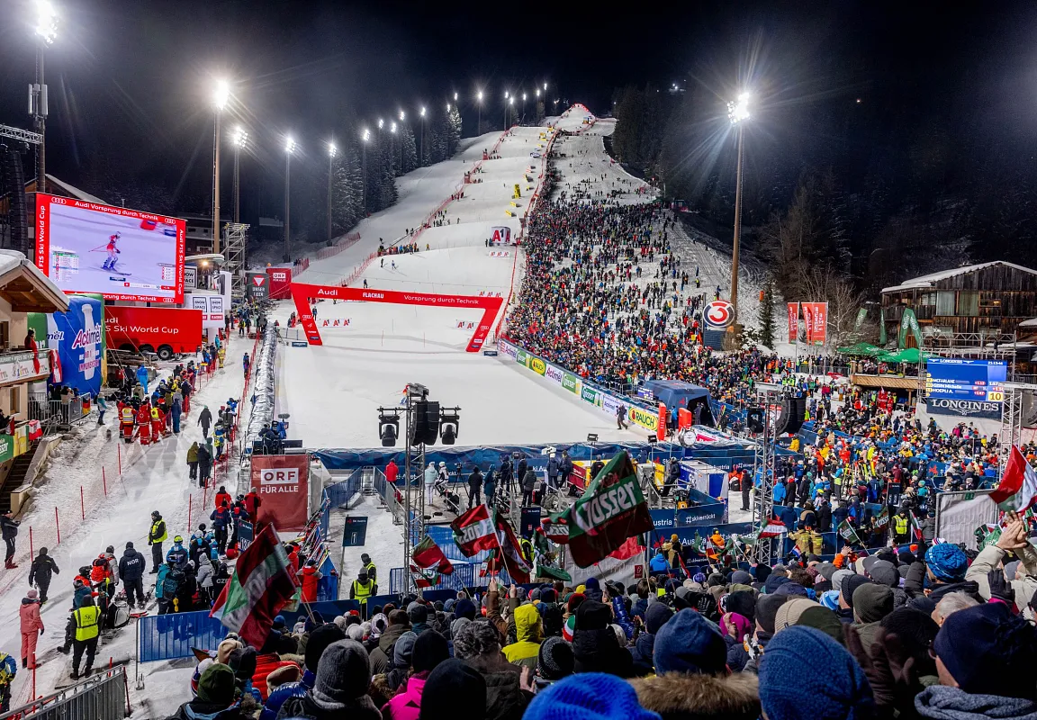 Ein beleuchteter Nachtskihang mit vielen Zuschauern, großen Bildschirmen und Skifahrern im Wettbewerb. Fahnen und Banner sind sichtbar, die Spannung liegt in der Luft.