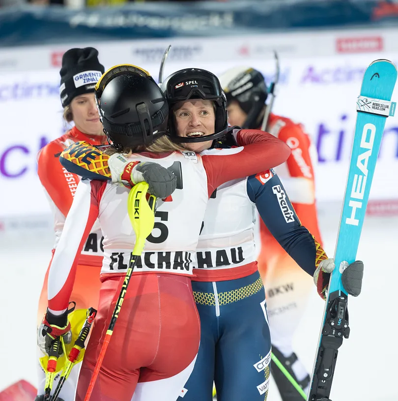 Two skiers in racing suits embrace warmly at a snowy event, surrounded by others. They hold ski poles and wear helmets and goggles, celebrating a race.