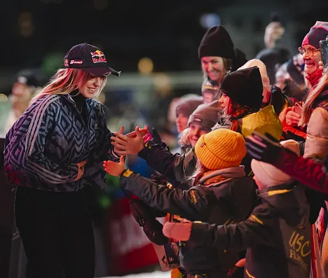 Eine Sportlerin in Winterkleidung begrüßt jubelnde Fans an einer Absperrung. Kinder und Erwachsene tragen warme Kleidung und Mützen, es herrscht fröhliche Stimmung.