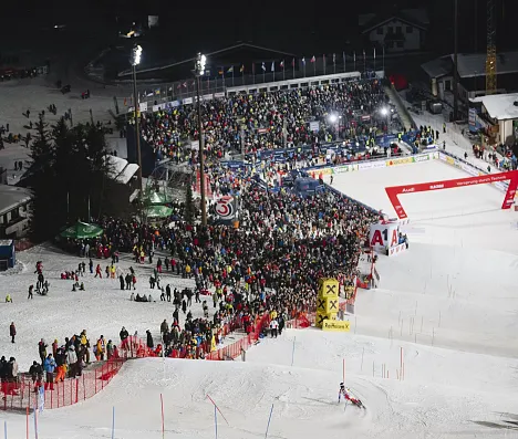 Skigebiet bei Nacht, beleuchtete Skipiste mit vielen Zuschauern. Eine Menschenmenge sammelt sich um den Zielbereich, um ein Skirennen zu verfolgen.